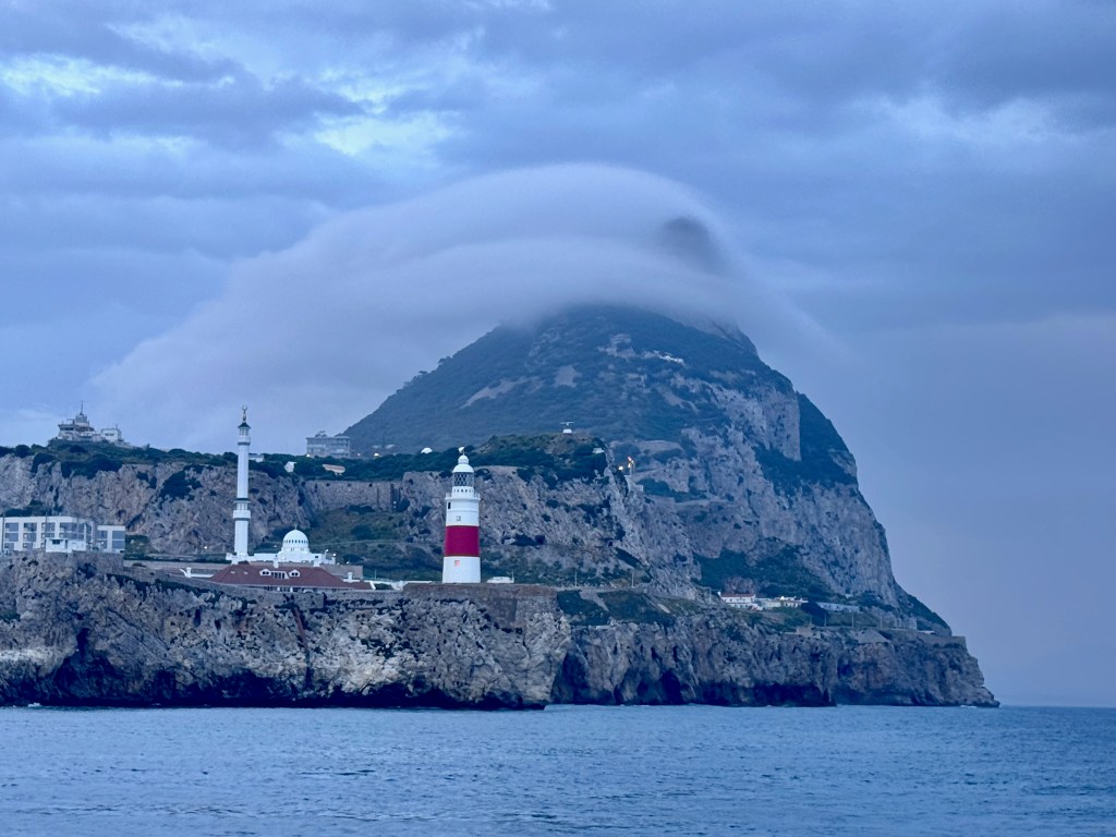 A view of a coastal landscape featuring a towering rock formation, partially covered by clouds, with lighthouses and buildings along the shore.