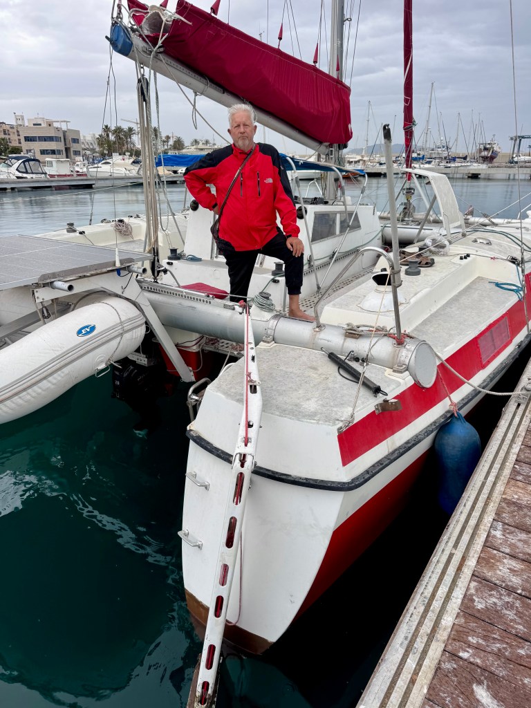A man in a red coat stands on the deck of a sailboat at a marina, with multiple boats docked in the background and cloudy skies above.