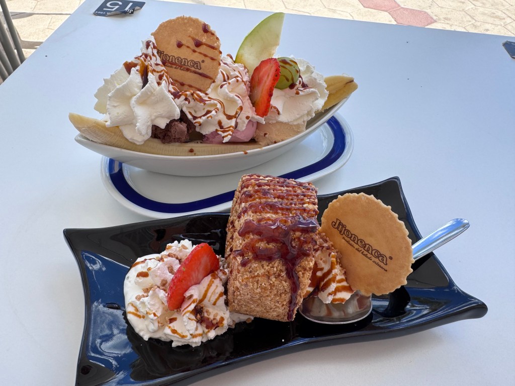 A delicious dessert display featuring a banana split with various ice creams, whipped cream, and a cookie on top, alongside a slice of cake drizzled with syrup, garnished with whipped cream and a strawberry.