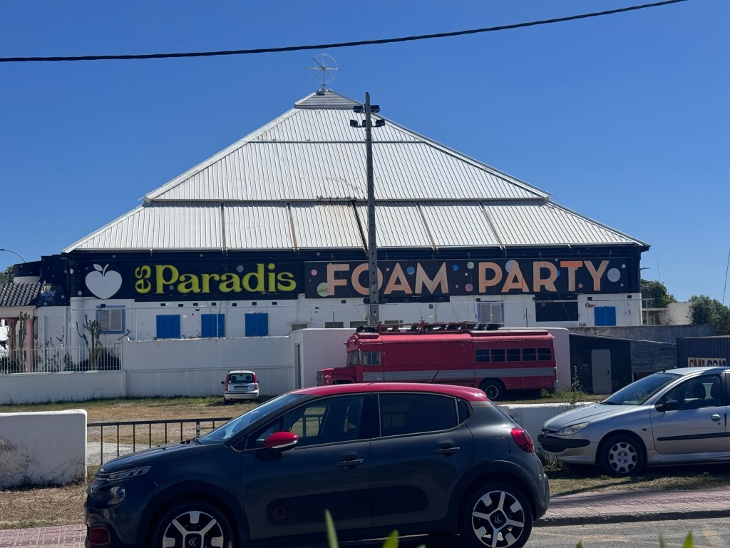 A large triangular-roofed building with a colorful sign that reads 'es Paradis FOAM PARTY.' Several parked cars are visible in the foreground.