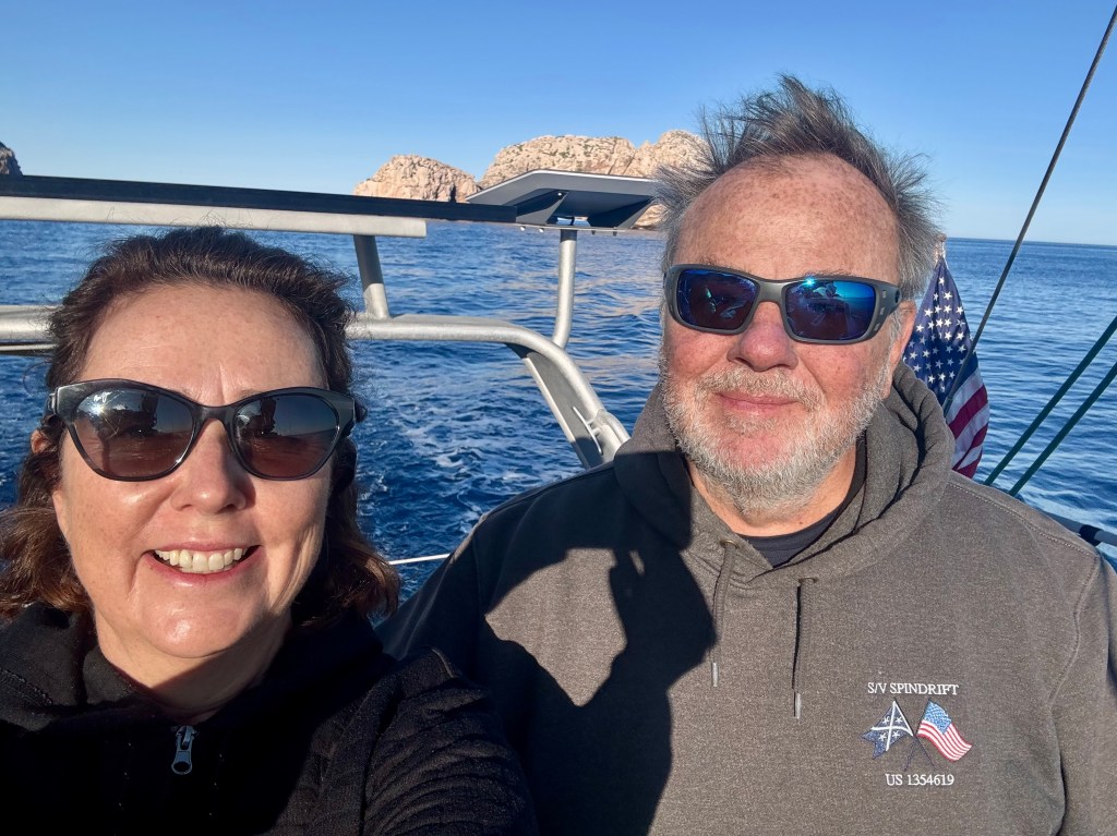 A smiling couple wearing sunglasses on a boat with rocky cliffs and clear blue water in the background.
