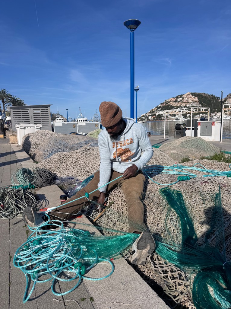 A person sitting on a dock surrounded by fishing nets and ropes, working on repairing equipment. The scene includes boats in the background and a clear blue sky.