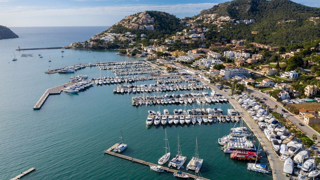 Aerial view of a marina filled with yachts, surrounded by hills and residential buildings, overlooking a calm sea.