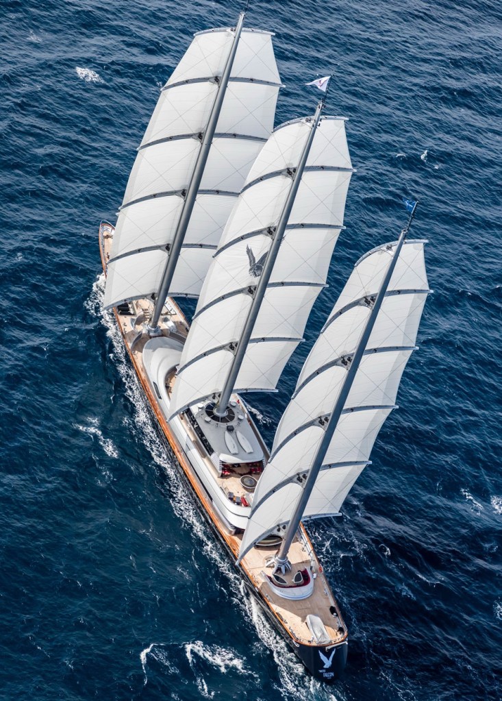 Aerial view of a large sailing yacht with multiple white sails navigating through blue ocean waters.
