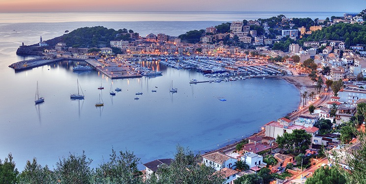 A scenic view of a coastal harbor at dusk, featuring a marina filled with boats, surrounded by buildings on a hillside and calm waters reflecting the twilight sky.