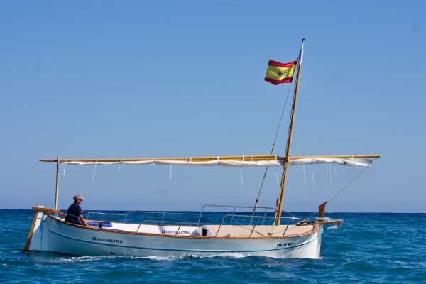 A traditional wooden boat sailing on clear blue water with a person on board and a Spanish flag flying.