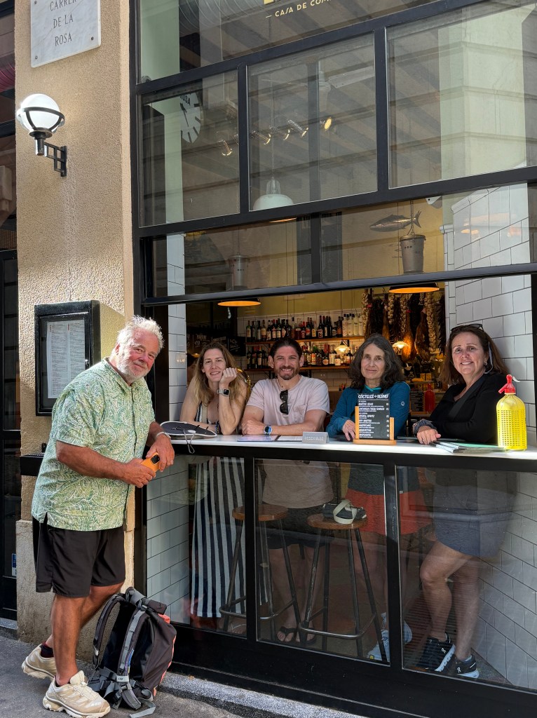 A group of five people, including a man in a patterned shirt and four others at a bar inside a restaurant, smiling at the camera. The restaurant features a backlit menu and various alcoholic beverages on display.