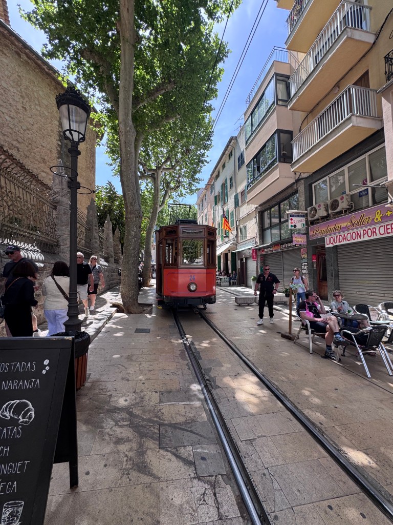 A quaint street view featuring a vintage tram in a pedestrian area, surrounded by trees and various shops. People are walking along the path, enjoying the sunny day.