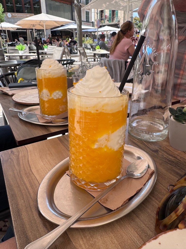 Two tall glasses of orange drink topped with whipped cream, placed on a wooden table in an outdoor café setting, with people dining in the background.