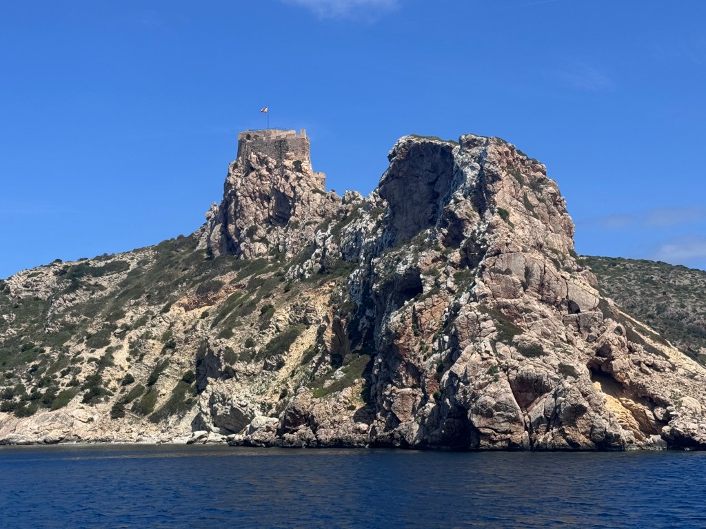 A rocky island with a historic castle perched on top, surrounded by clear blue water and a blue sky.