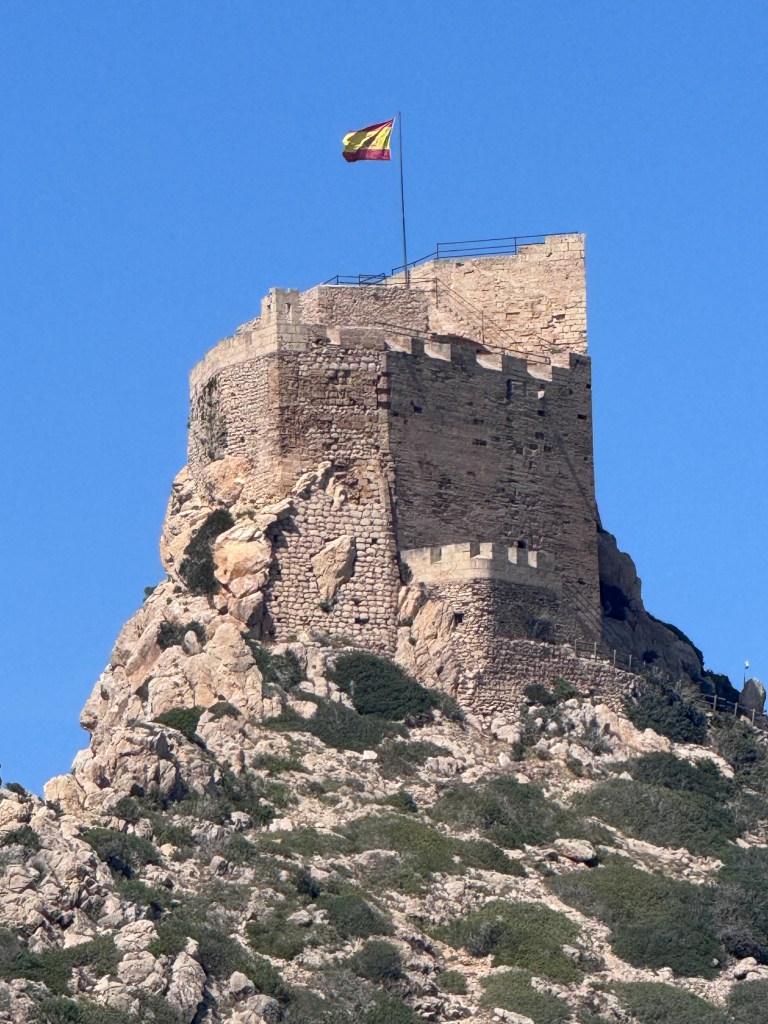 A historic stone castle perched on a rocky hilltop, with a flag flying from its highest point under a clear blue sky.