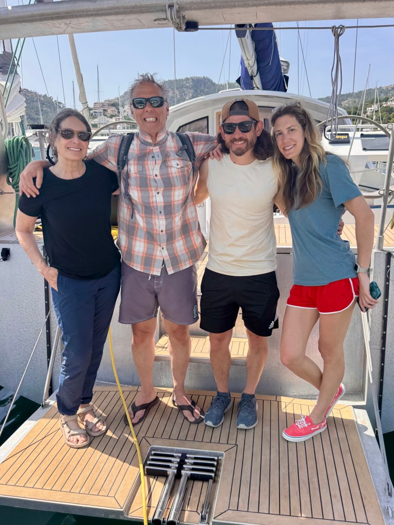 Four people posing on a boat deck smiling, with the sea and hills in the background.