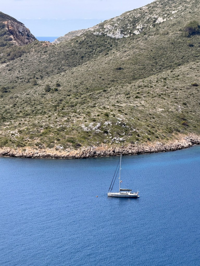 A sailboat anchored in calm blue waters near a rocky coastline, surrounded by lush green hills.