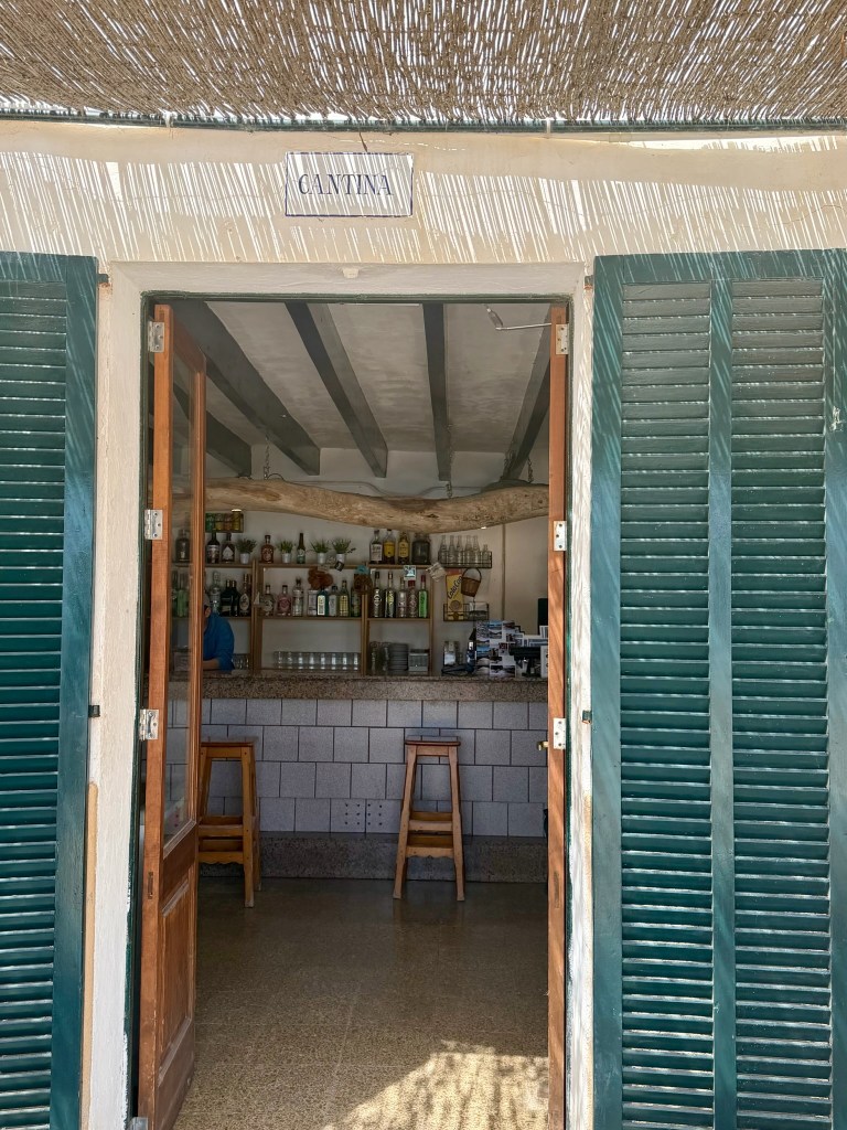 Entrance to a cantina with open wooden doors and slatted green shutters, showcasing a bar area stocked with bottles.