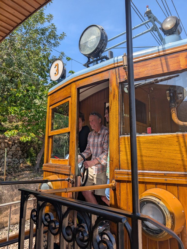 A vintage wooden tram with the number 3 displayed on the front, featuring a driver at the controls and a few passengers visible inside, set against a backdrop of greenery and blue sky.
